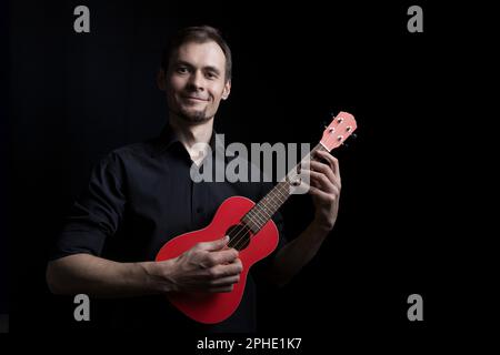Handsome smiling low key man portrait playing on fun red ukulele guitar over black background Stock Photo