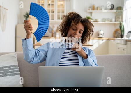 Exhausted overheated African American girl freelancer waving hand fan and working on laptop at home Stock Photo