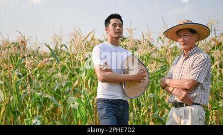 A successful millennial Asian male farm owner or supplier and an elderly farmer are standing with arms crossed in a cornfield. Stock Photo