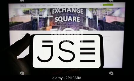 Person holding smartphone with logo of JSE Limited (Johannesburg Stock Exchange) on screen in front of website. Focus on phone display. Stock Photo