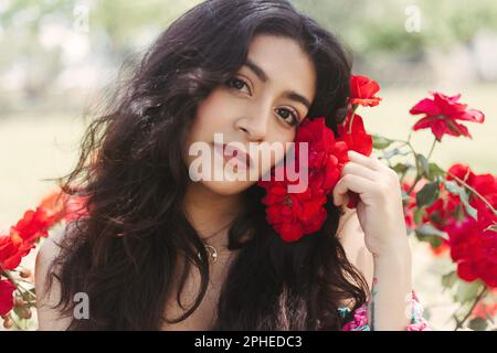 Pensive young female with long dark hair holding red flowers in hand and looking at camera while standing in garden against blurred background of natu Stock Photo