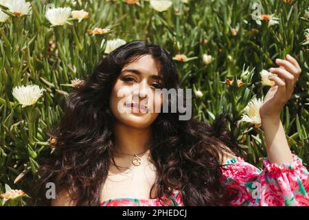 From above of smiling young female with long dark hair and raised hand looking at camera while lying on green plants with white blooming flowers in su Stock Photo