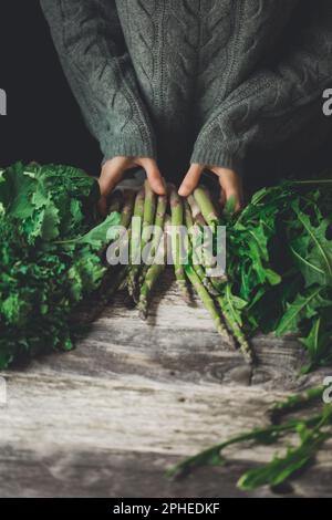 Unrecognizable person in gray sweater standing at wooden table with fresh asparagus stems Stock Photo