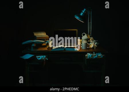Stack of various books and supplies placed on wooden table with mess and cups near glowing lamp in dark room Stock Photo