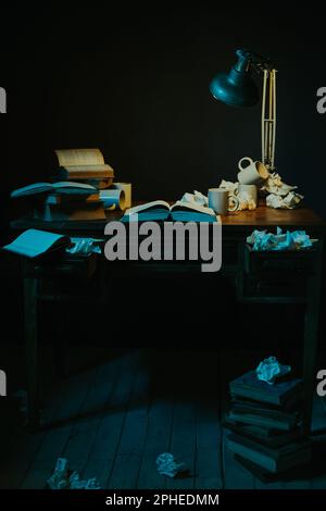 Stack of various books and supplies placed on wooden table with mess and cups near glowing lamp in dark room Stock Photo