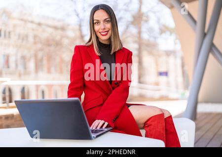 Happy young woman in dark blonde hair and red lipstick sitting at table and looking at camera screen against blurred background Stock Photo