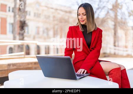 Happy young woman in dark blonde hair and red lipstick sitting at table and looking at laptop screen against blurred background Stock Photo