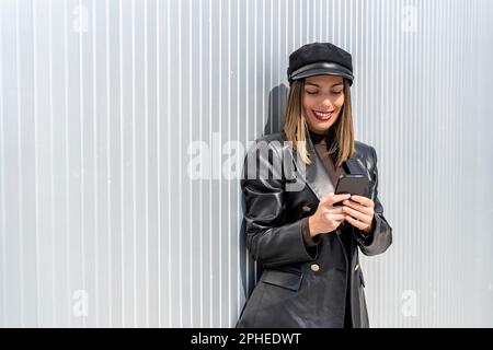 Young female in black leather jacket and cap smiling while browsing cellphone against gray wall in street Stock Photo