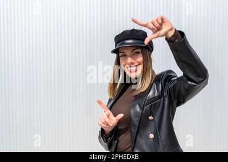 Positive young female in leather jacket and cap looking at camera while showing frame gesture against gray wall Stock Photo
