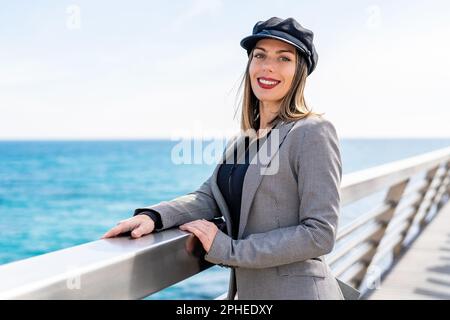 Young smiling woman in gray jacket and black hat pointing on camera while standing near fence in background of sea Stock Photo