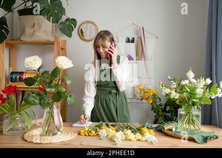 Cheerful woman wearing green overall and making notes while talking on smartphone and receiving order in florist shop with various flowers Stock Photo