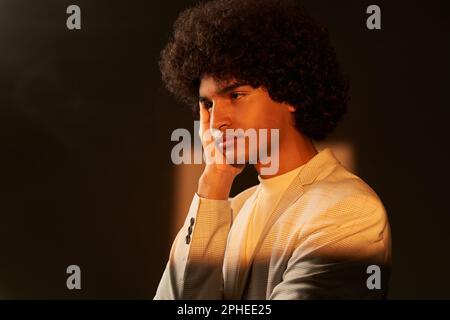 Side view of young ethnic thoughtful Afro haired male model wearing checkered jacket and touching cheek while standing in darkness with rectangular or Stock Photo