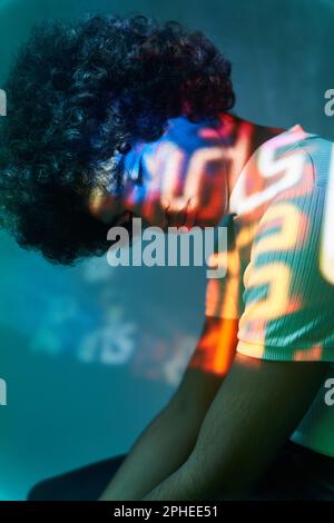 Ethnic man with Afro hair leaning head forward while sitting against geometric illumination from projector in dark studio Stock Photo