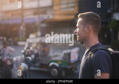 Portrait of tourist with backpack walking at street. Side view of man against tuk tuk on road. Bangkok, Thailand Stock Photo
