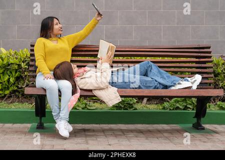 Woman reading on a bench next to a friends that is taking a selfie Stock Photo