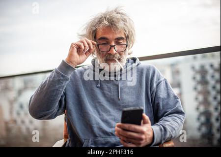 Handsome senior man with grey hair and eyeglasses sitting on terrace, using cigarette and smartphone, communicating, having fun and enjoying Stock Photo