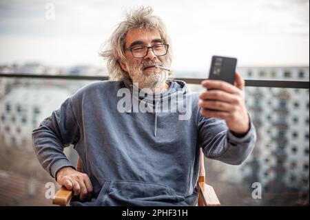Handsome senior man with grey hair and eyeglasses sitting on terrace, using cigarette and smartphone, communicating, having fun and enjoying Stock Photo