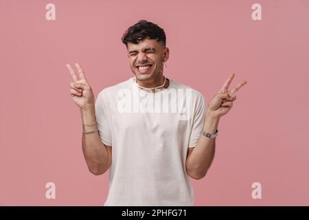 Young handsome stylish smiling man with closed eyes and raised hands showing victory gesture with fingers , while standing over isolated pink backgrou Stock Photo