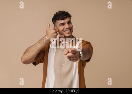 Young handsome stylish smiling man doing phone gesture wit hand and fingers, pointing and looking at camera, while standing over isolated brown backgr Stock Photo