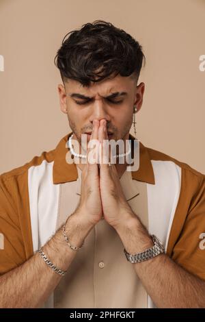 Young handsome stylish frowning man with closed and foldedhands doing pray gesture, while standing over isolated brown background Stock Photo