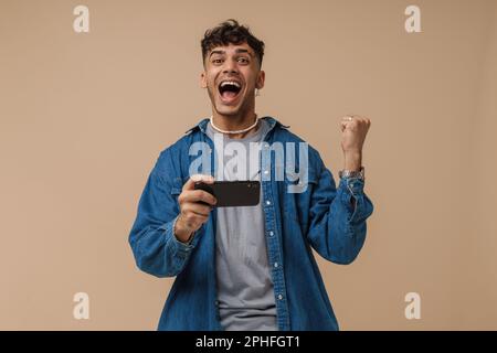Young handsome stylish enthusiastic man holding phone doing winner gesture with raised fist and opened mouth and looking at camera , while standing ov Stock Photo