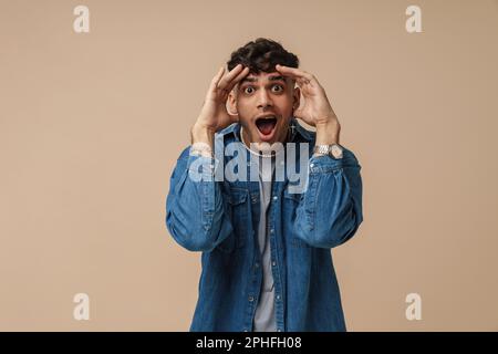 Young handsome stylish shocked man with opened mouth and wide opened eyes touching his head and looking at camera, while standing over isolated brown Stock Photo
