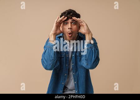 Young handsome stylish shocked man with opened mouth and wide opened eyes touching his head and looking at camera, while standing over isolated brown Stock Photo