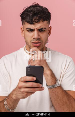 Young hadsome stylish thoughtful man looking at phone in his hand and touching his chin, while standing over isolated pink background Stock Photo