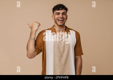 Brunette young man laughing and pointing finger aside isolated over beige background Stock Photo