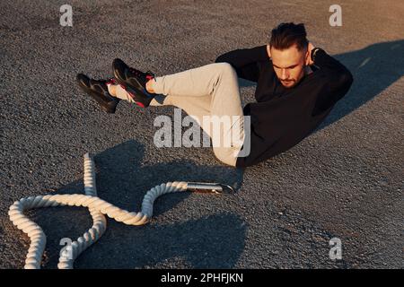 Doing abs. Man in sportswear have fitness day on the road at evening time Stock Photo