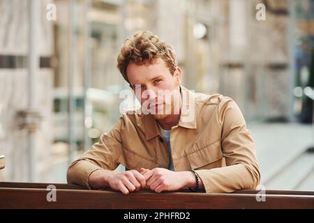 Elegant young man in formal classy clothes leaning on wooden fence outdoors in the city Stock Photo