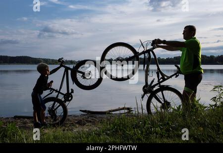 Silhouettes of an adult man and a boy with bicycles in helmets near the lake. Family vacation, cycling, travel, healthy lifestyle. leisure. father's d Stock Photo