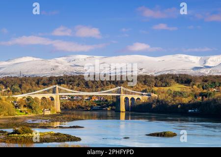 Scenic view to the Menai Suspension bridge crossing the Menai Strait with snow on the mountains in winter. Menai Bridge, Isle of Anglesey, Wales, UK Stock Photo
