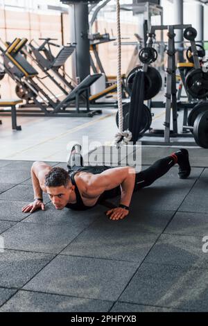Doing push-ups. Young sportive strong man in black wear have workout day in gym Stock Photo