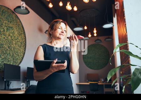 Businesswoman having business call working on tablet in office. Mature busy woman using touch pad computer standing by window in modern interior. Fema Stock Photo