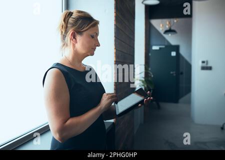 Businesswoman working on tablet in office. Mature woman using touch pad computer standing by window in modern interior. Female manager focused on work Stock Photo