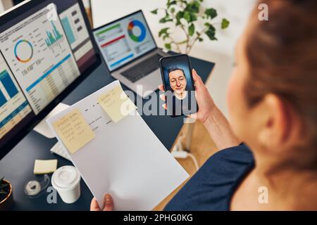 Businesswoman having video chat on mobile phone with her colleague. Businesswoman working with data on charts, graphs and diagrams on computer screen. Stock Photo