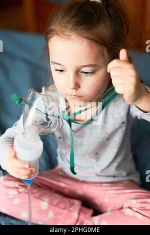 Sick little girl having medical inhalation treatment with nebuliser. Child holding breathing mask sitting in bed Stock Photo
