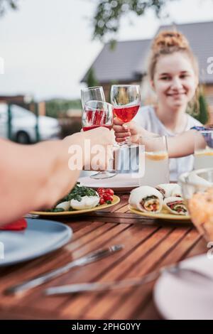 Family making toast during summer outdoor dinner in a home garden. Close up of hands holding wine glasses with red wine over table with dishes Stock Photo