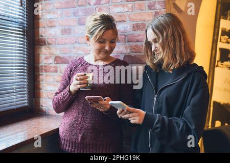 Smiling women using smartphones drinking coffee charging mobile phone relaxing in office while taking a break Stock Photo