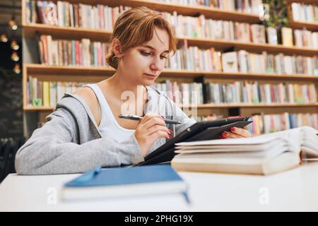 Student learning in university library. Young woman writing essay and making notes using computer tablet. Focused student studying for college exams. Stock Photo