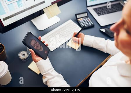 Woman entrepreneur having business video chat on smartphone. Businesswoman making notes while talking to her co-worker. Woman holding smartphone sitti Stock Photo