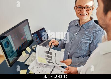 Woman entrepreneur discussing financial data with her colleague standing at desk in office. Happy positive businesswoman working with charts and table Stock Photo