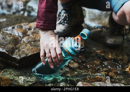 Woman taking pure water to bottle from mountain stream during trekking in mountains. Hiker crouching on rocks, filling bottle up with cold mountain wa Stock Photo