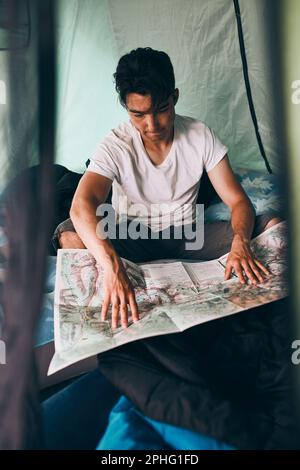 Young man looking at a map of mountain trails planning next trip on summer vacation sitting in a tent on camping. Concept of camp life Stock Photo