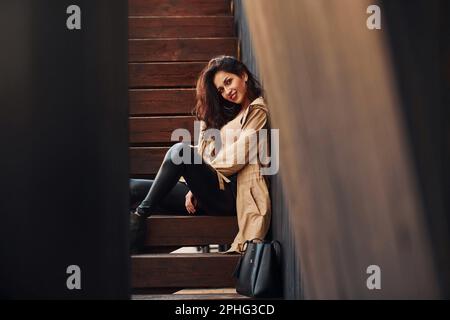 Cheerful brunette in black leggings sitting on wooden stairs and smiling Stock Photo