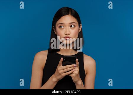Young beautiful asian woman with puckered lips holding phone and looking aside, while standing over blue background Stock Photo