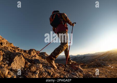 Hiker with backpack walk uphill in mountains. Active tourist silhouette goes with hiking poles at sunset Stock Photo