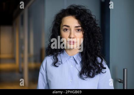 Portrait of a young Muslim woman with curly black hair. Students, businesswomen, freelancers standing in the office and looking at the camera. Stock Photo