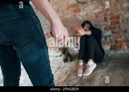 Violent man standing with knife in hand and threatens girl that sits on the floor in abandoned building Stock Photo